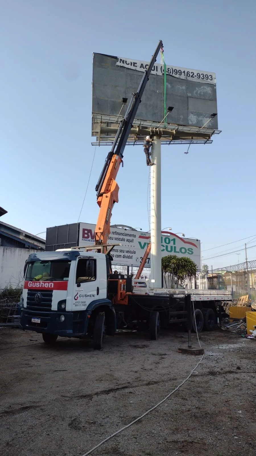 Caminhão munck com a lança estendida instalando a placa de um painel publicitário no alto de um mastro.
