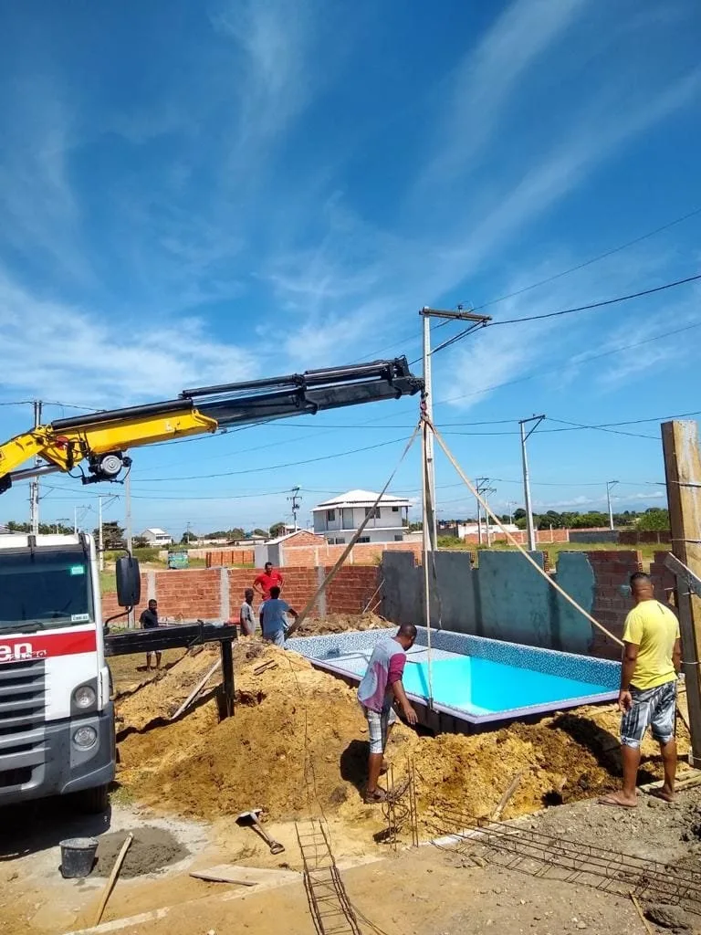 Caminhão munck baixando uma piscina de fibra dentro da escavação em um terreno.