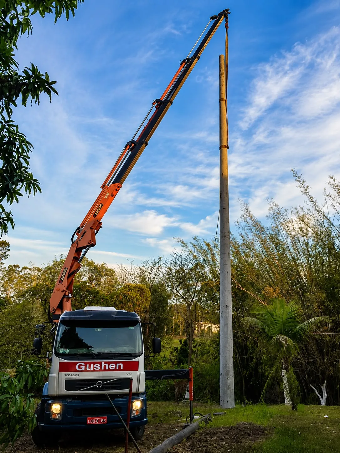 Caminhão munck com a lança totalmente estendida implantando um poste de concreto.
