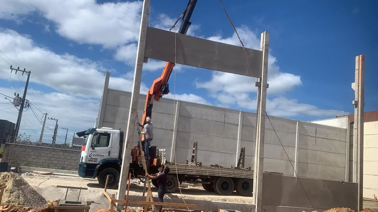 Caminhão munck da Gushen posicionando uma viga pré-moldada de concreto entre pilares, com dois trabalhadores na montagem.