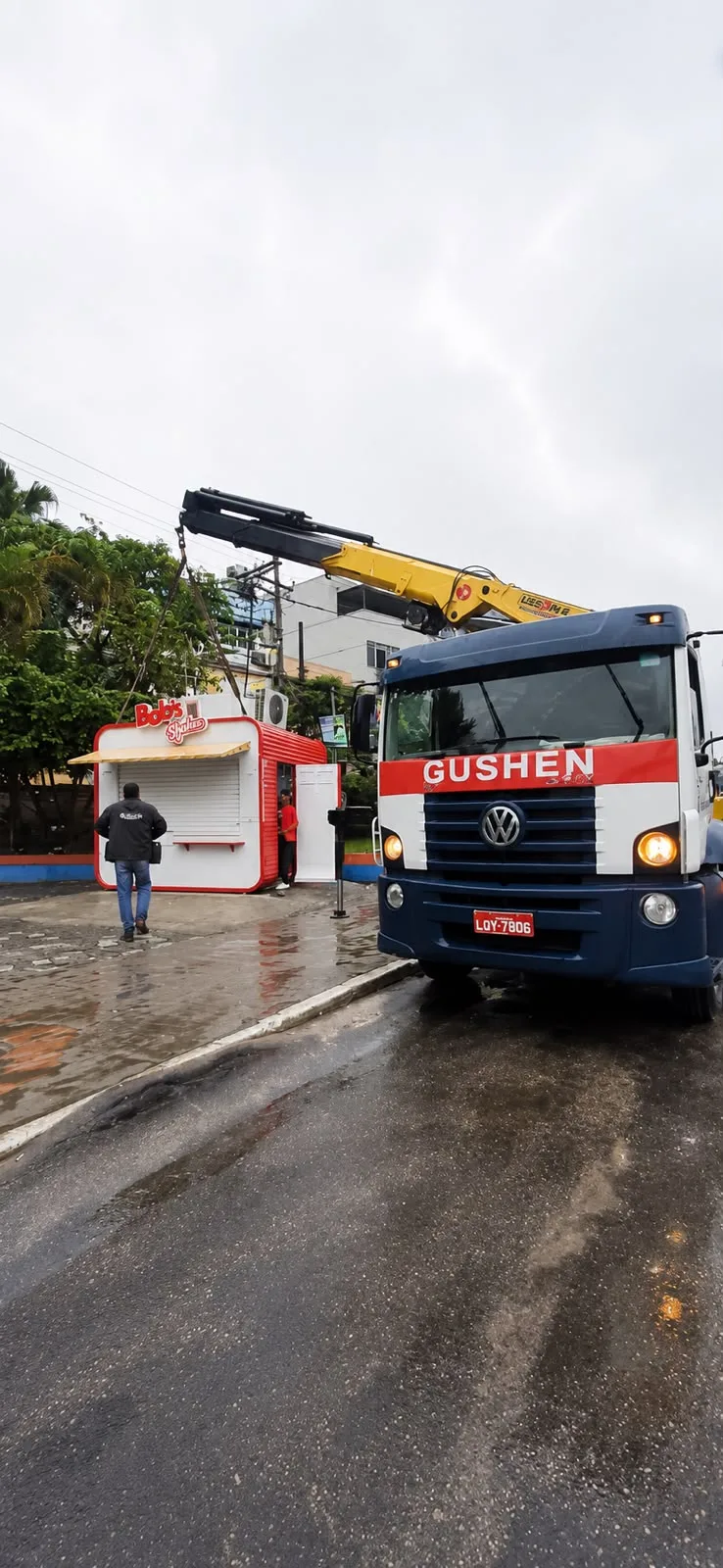Caminhão munck da Gushen com braço amarelo em rua molhada, ao lado de um quiosque recém-instalado.