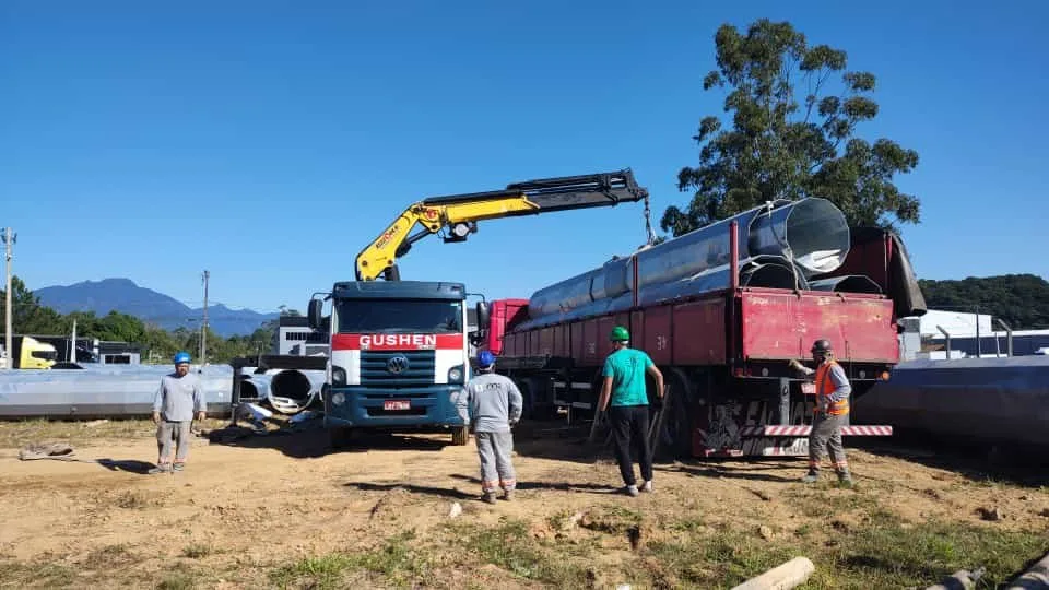 Caminhão munck da Gushen descarregando tubos de grande diâmetro de uma carreta, com a equipe acompanhando.
